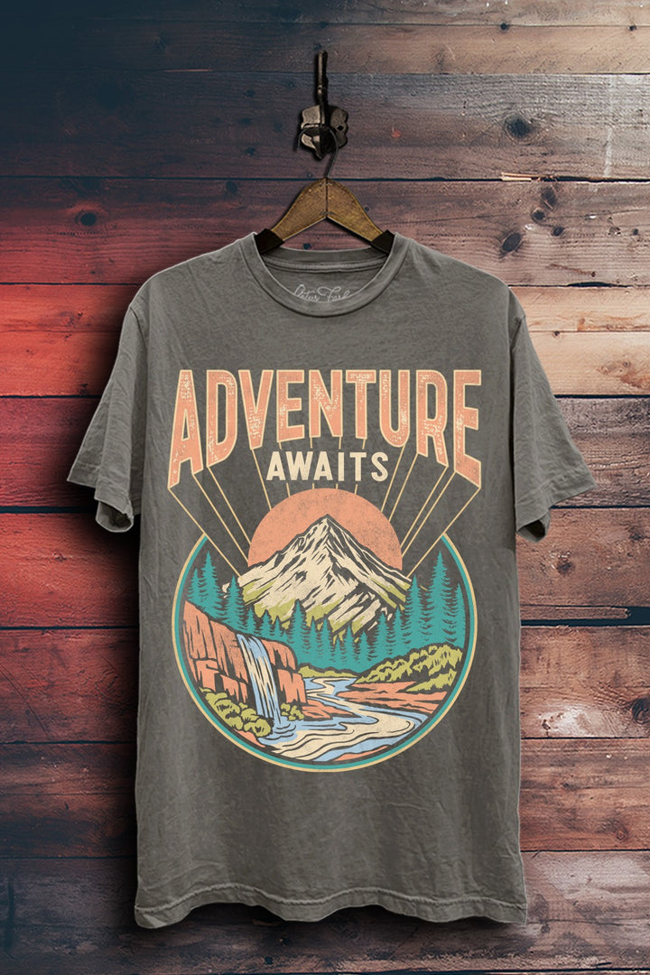 Stone gray mineral-washed plus-size boyfriend tee featuring Adventure Awaits lettering above a mountain, pine forest, waterfall, and winding river.