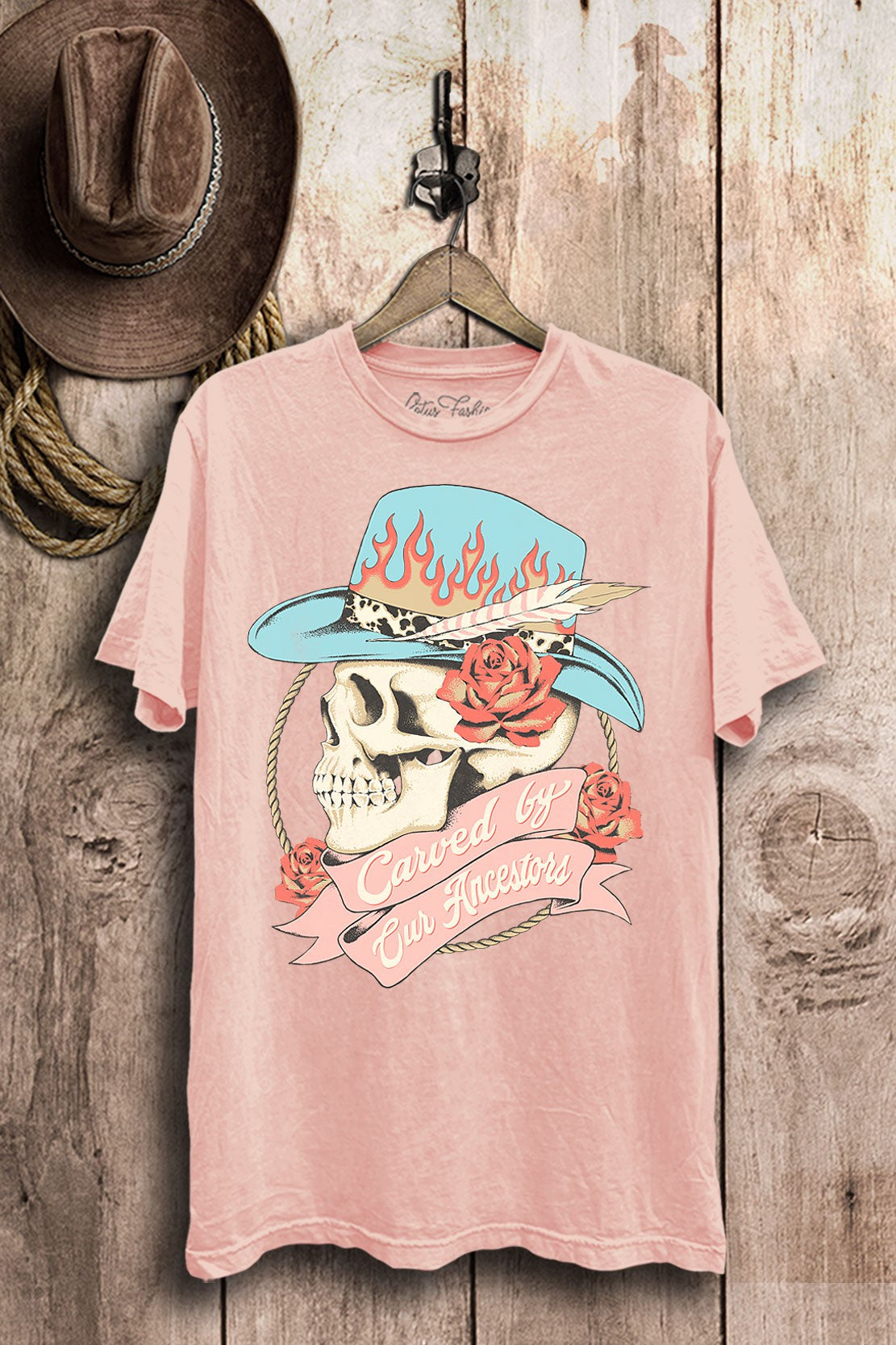 Light-pink mineral-wash boyfriend tee with a cowgirl skull, turquoise flame hat, coral roses, and Carved by Our Ancestors banner.