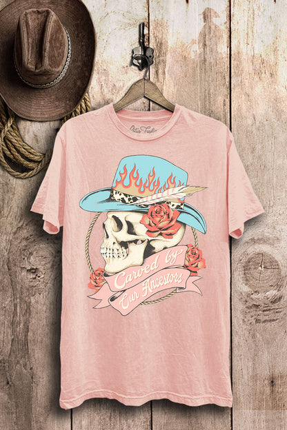 Light-pink mineral-wash boyfriend tee with a cowgirl skull, turquoise flame hat, coral roses, and Carved by Our Ancestors banner.