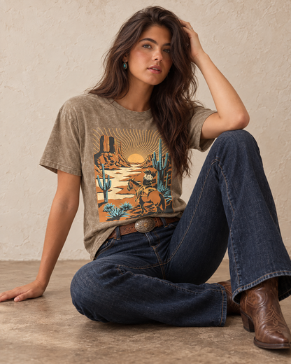 Mocha mineral-washed boyfriend tee featuring a cowboy riding through a sunset desert with mesas, tall cacti, and turquoise-colored plants.