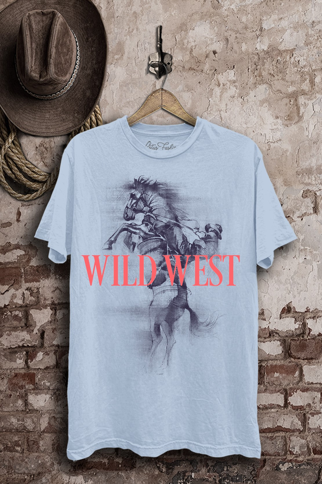 Light-blue mineral-wash boyfriend tee featuring a cowboy riding a bucking horse with bold coral Wild West lettering.